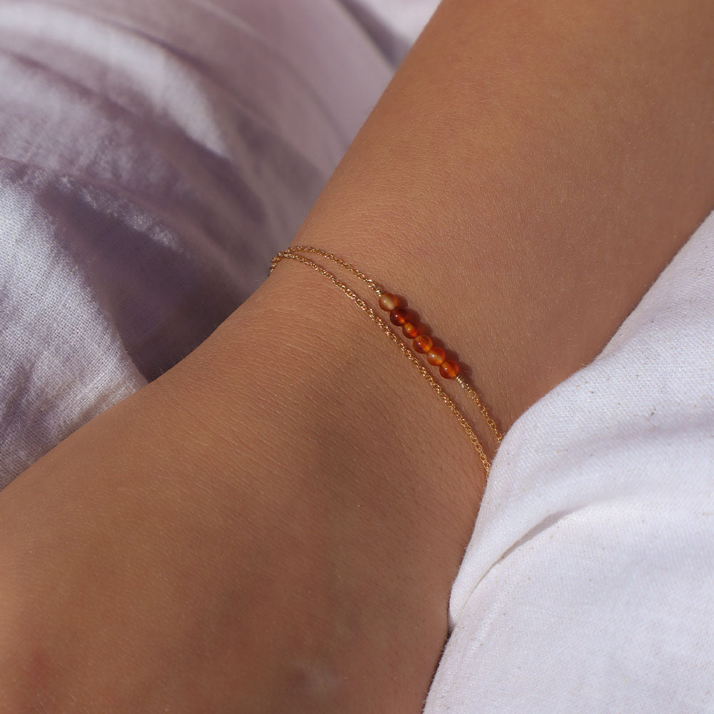 Close-up of a wrist wearing a gold bracelet with orange beads against a blurred background.