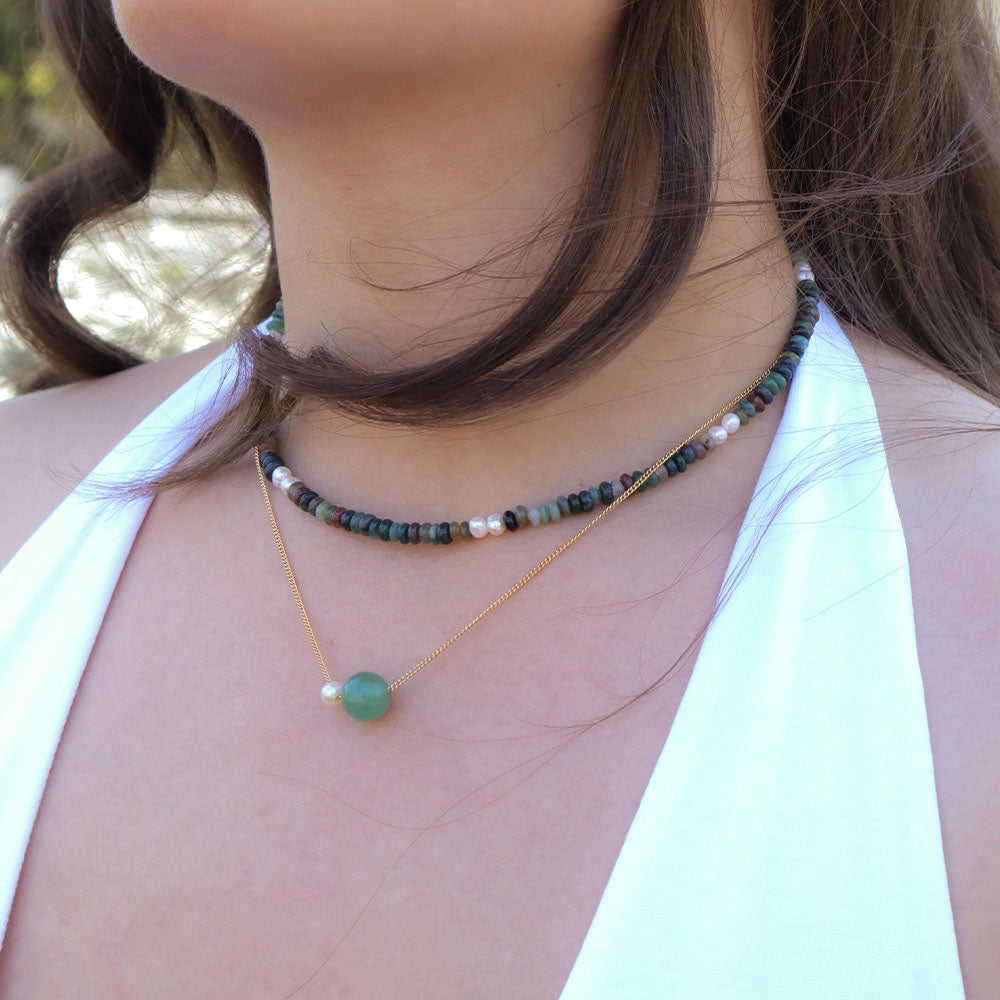 Close-up of a person wearing two necklaces with beads, one green and one gold, against a blurred natural background.