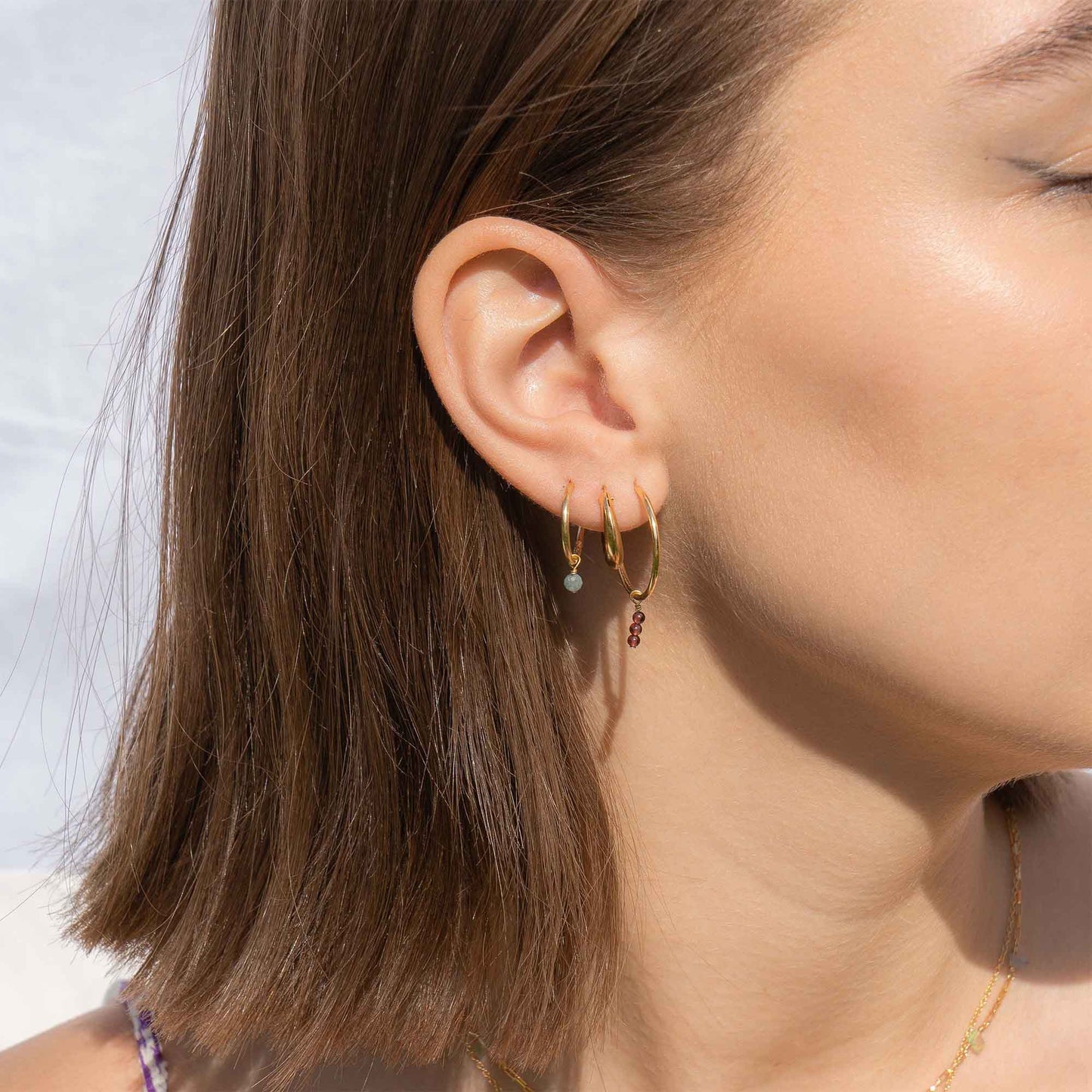 Close-up of a person wearing gold hoop earrings with small gemstones against a neutral background