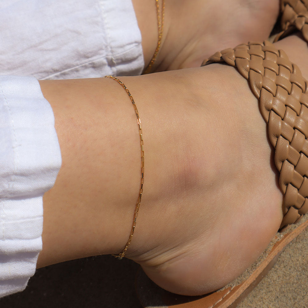 Close-up of a foot wearing a gold anklet with a woven sandal on a blurred background