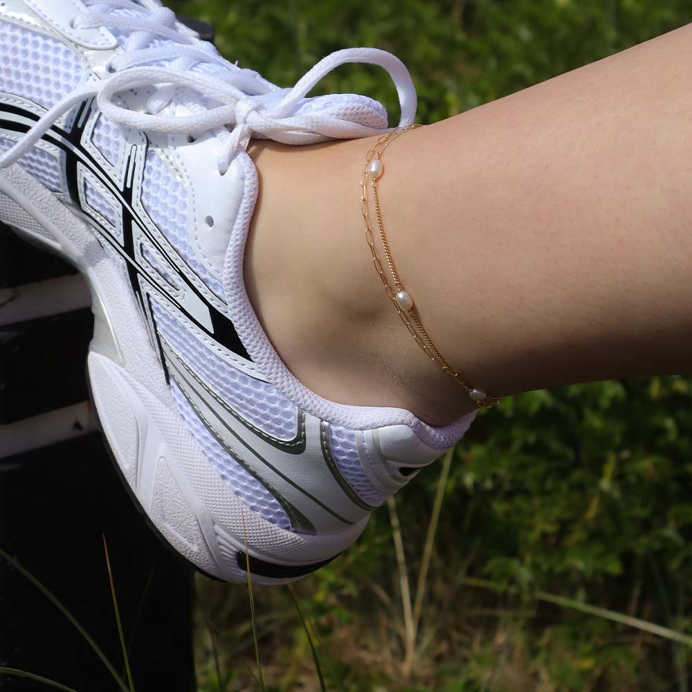 White athletic shoe with black accents on a person's foot, wearing a gold anklet with pearls, against a blurred natural background.