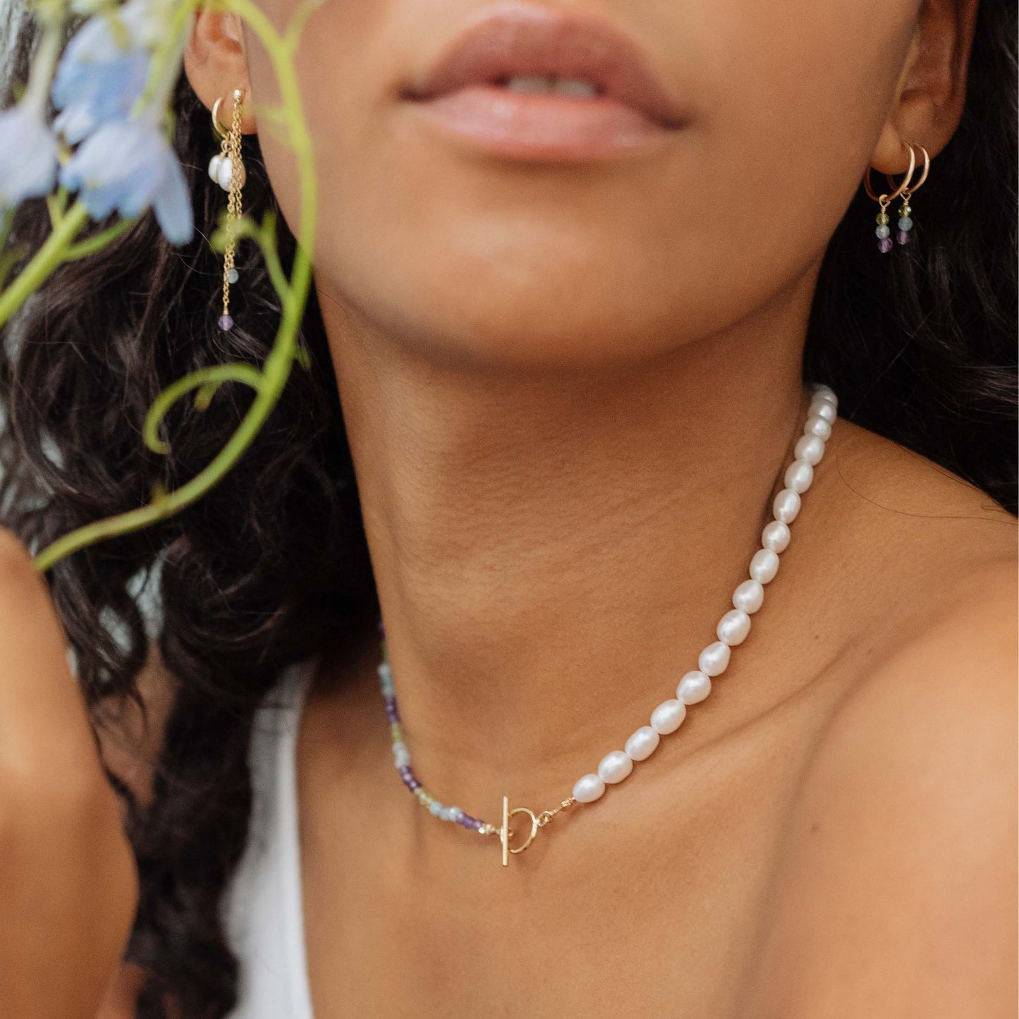 Close-up of a person wearing a pearl necklace with a gold clasp, surrounded by greenery.