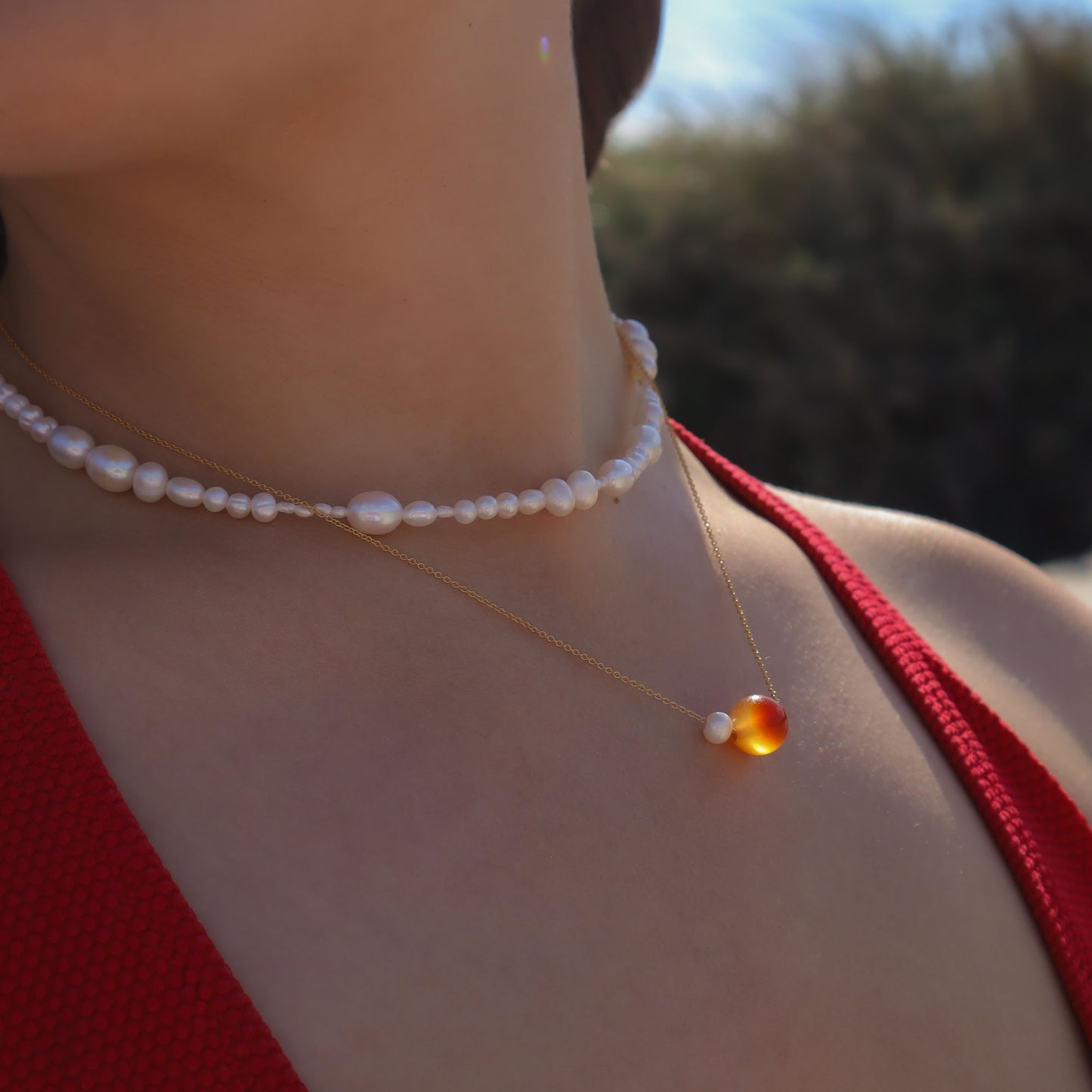 Close-up of a person wearing a pearl necklace and a red top with a blurred natural background