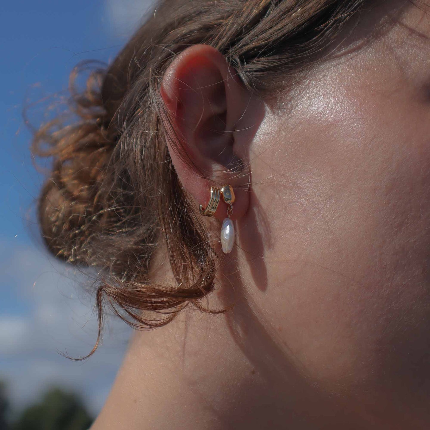 Close-up of an ear wearing a gold earring with a pearl, against a blurred natural background.