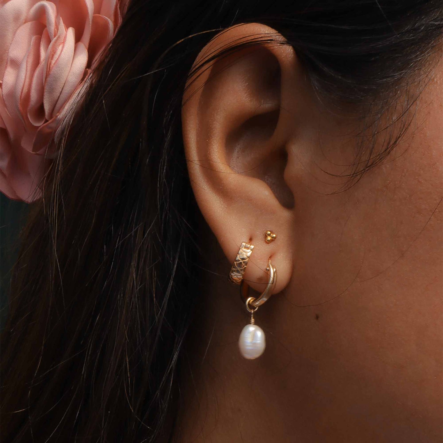 Close-up of an ear with gold earrings and a pearl pendant, with dark hair and a pink flower in the background.