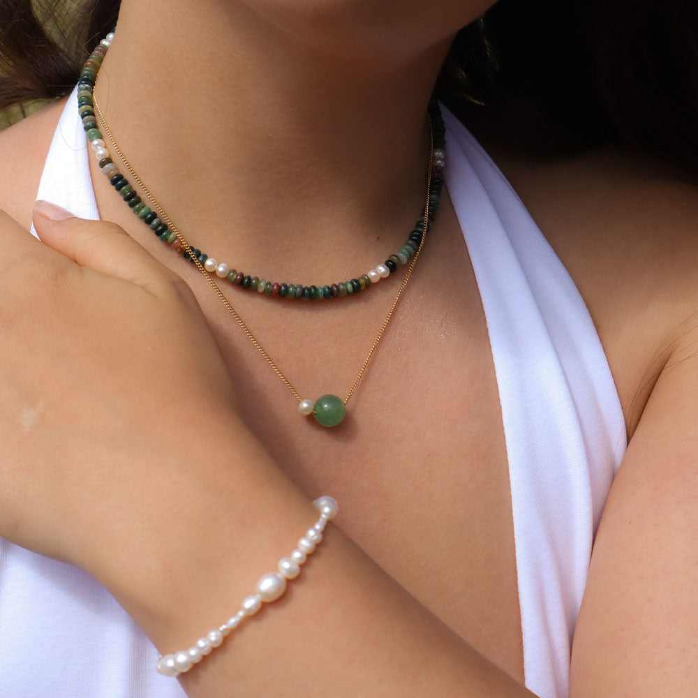 Close-up of a person wearing a green beaded necklace and a gold necklace with a green pendant, with a white top visible.