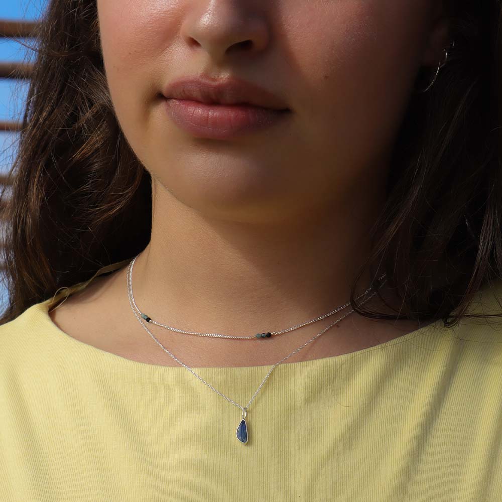 Close-up of a person wearing a necklace with a pendant, set against a blurred outdoor background.