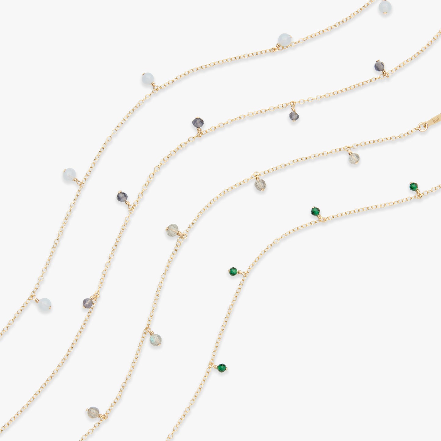 Three delicate gold chain necklaces with small, spaced gemstone beads in white, gray, green, and clear, arranged diagonally on a light gray background.