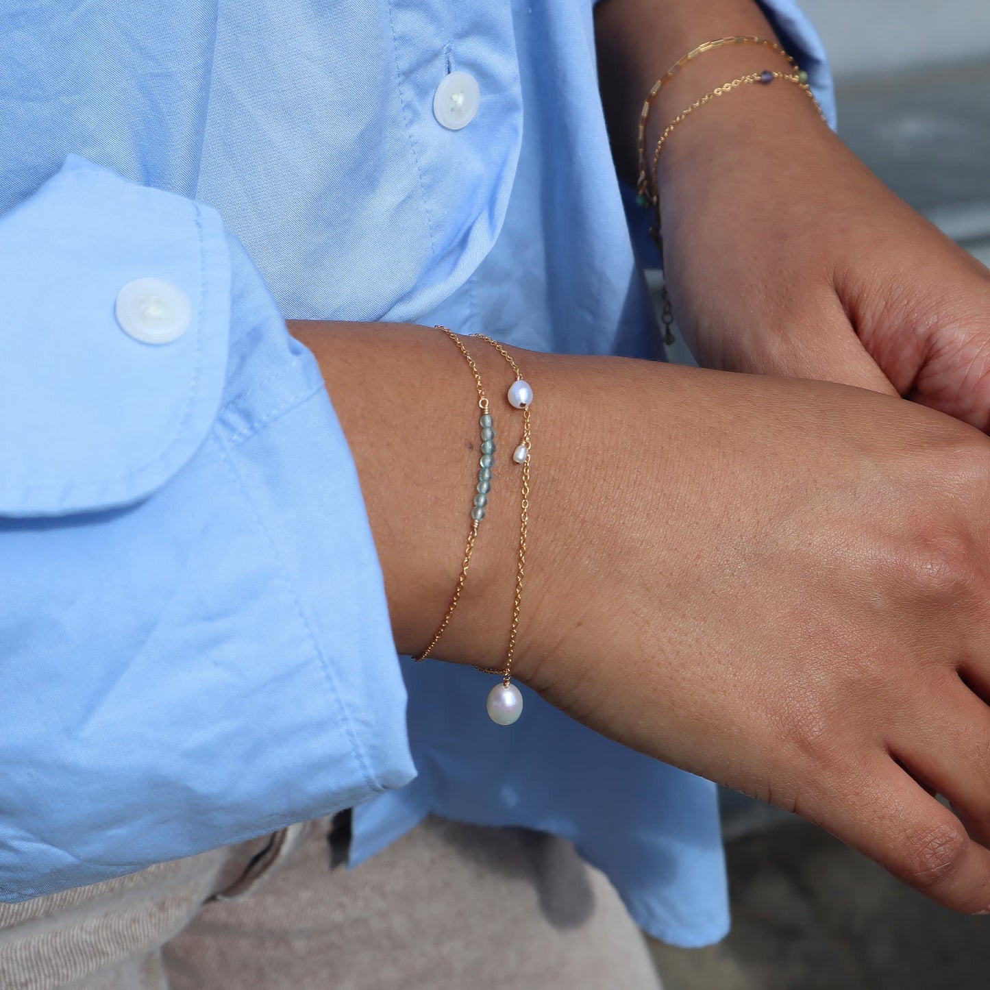 Close-up of a person wearing a gold bracelet with a pearl charm, against a blurred background.
