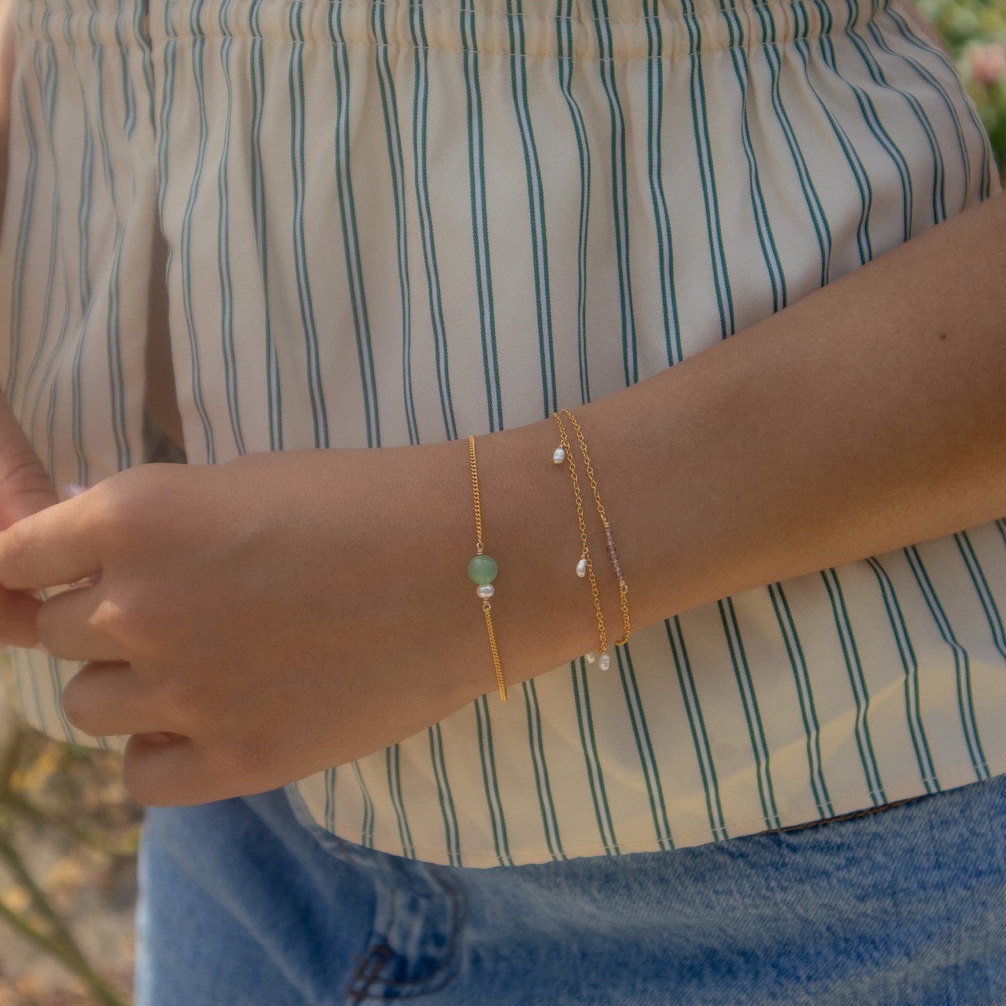 A close-up of a person's wrist wearing two delicate gold chain bracelets, one with a small light green bead and the other with tiny white beads, worn over a cream and blue striped top.