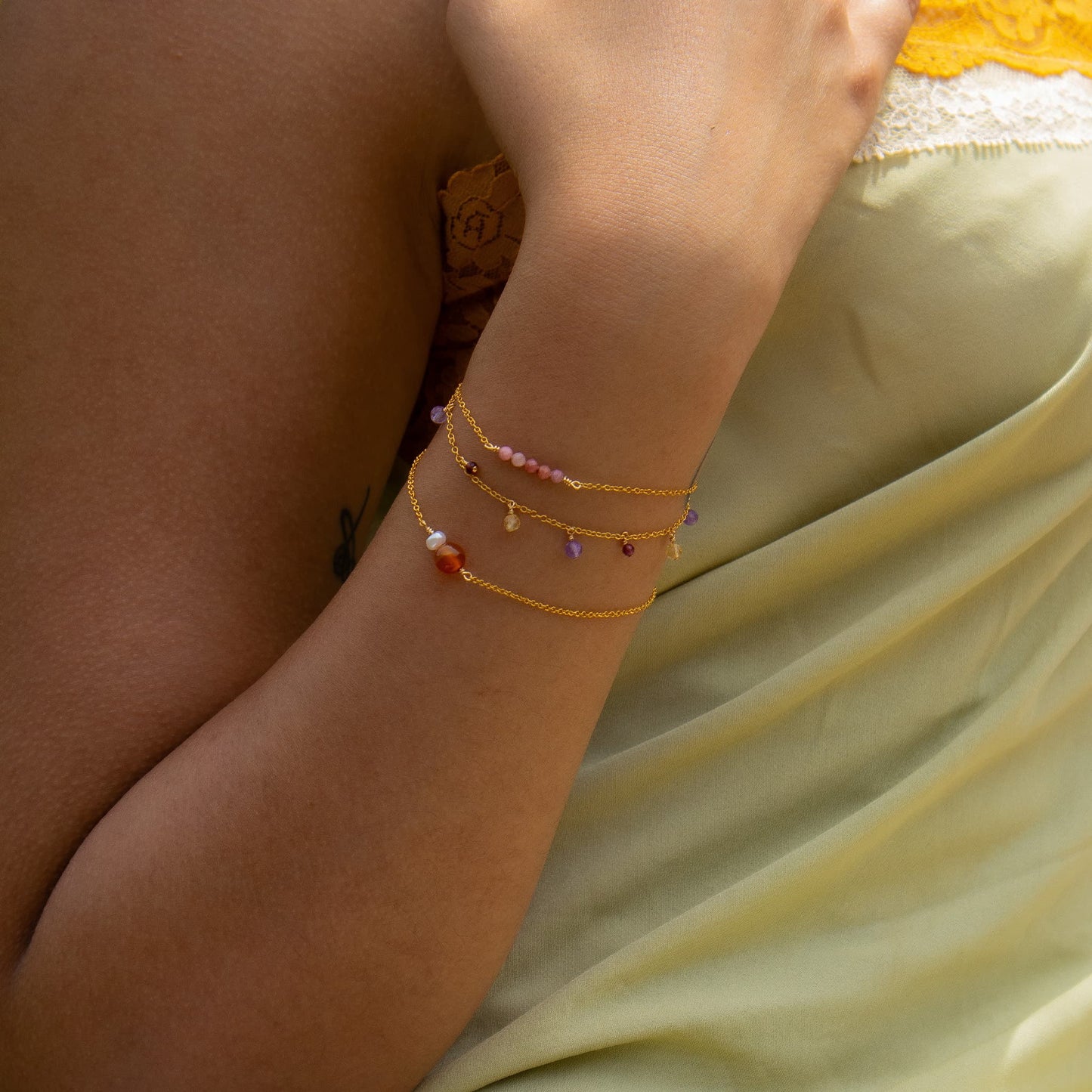A close-up of a person's wrist wearing three delicate gold chain bracelets adorned with small colorful beads and charms, resting against a pale yellow fabric background.