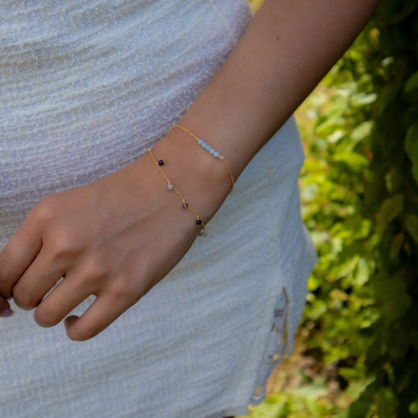 A close-up of a person's wrist wearing two delicate gold chain bracelets, one with small black beads and the other with light blue beads, set against a soft white textured top and a blurred green outdoor background.