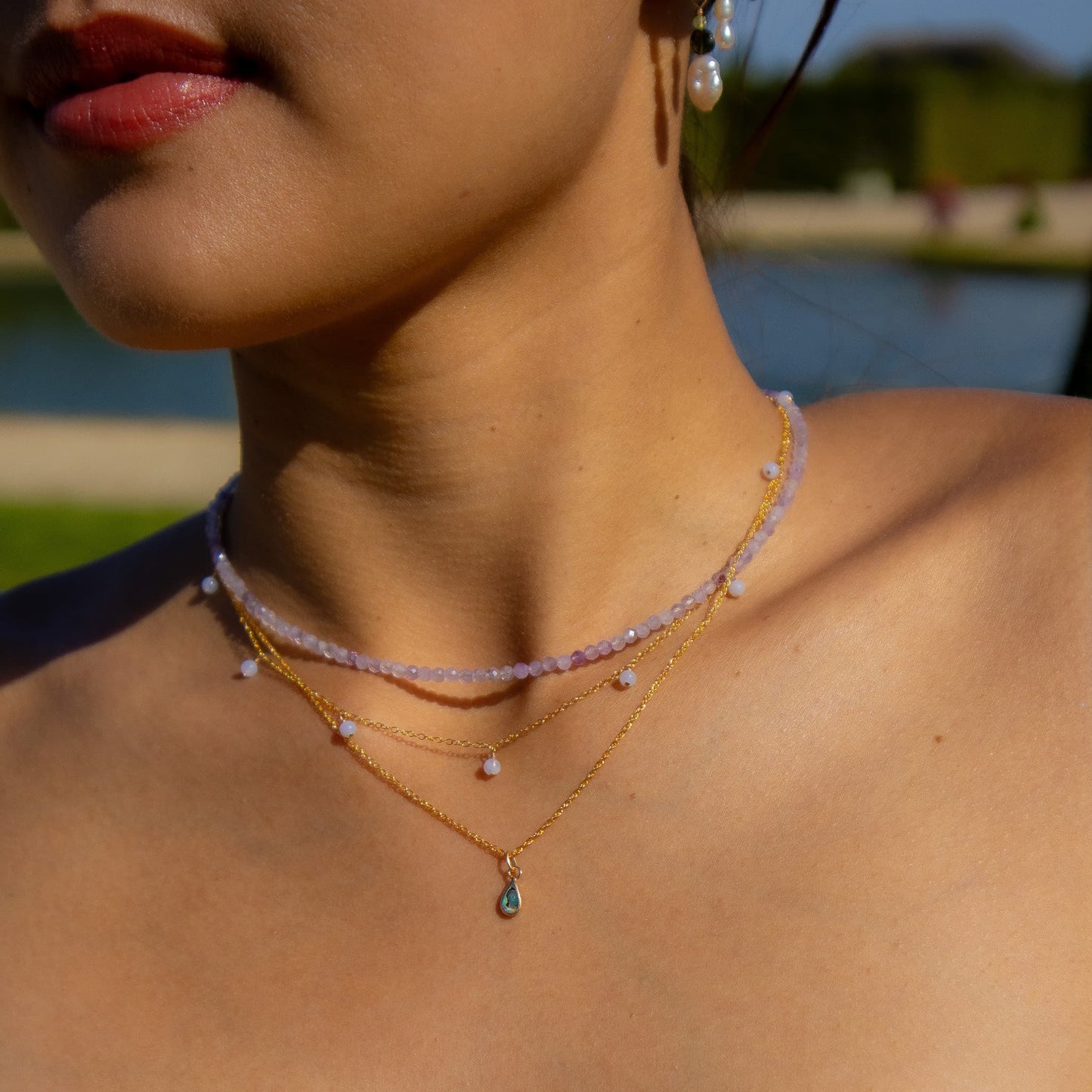 A close-up lifestyle shot of a model wearing a layered necklace set outdoors. The set features a light pink beaded choker, a delicate gold chain with small white pearl-like beads, and a gold chain with a small green teardrop pendant.