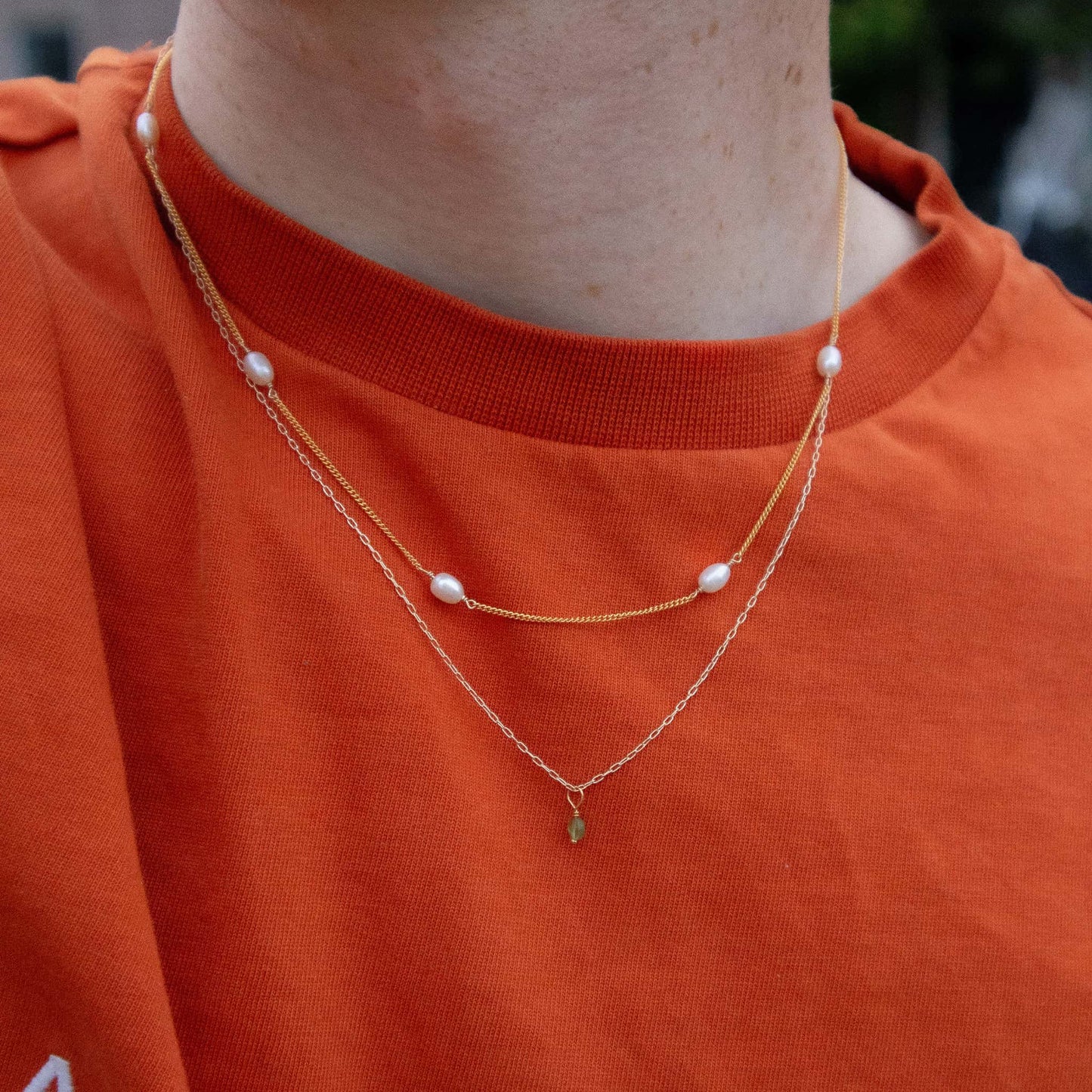 Person wearing a gold necklace with pearl-like beads on an orange shirt.