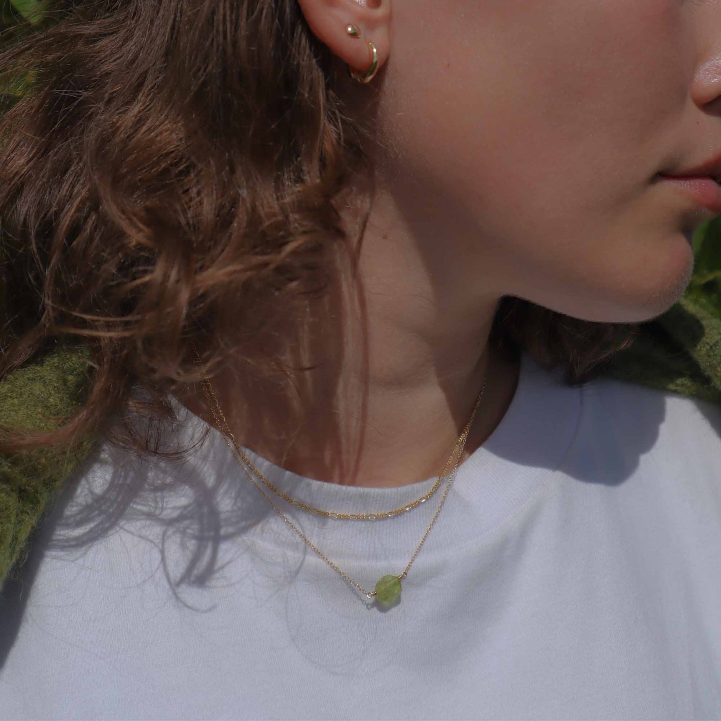 Close-up of a person wearing gold earrings and a necklace with a green pendant on a white background.