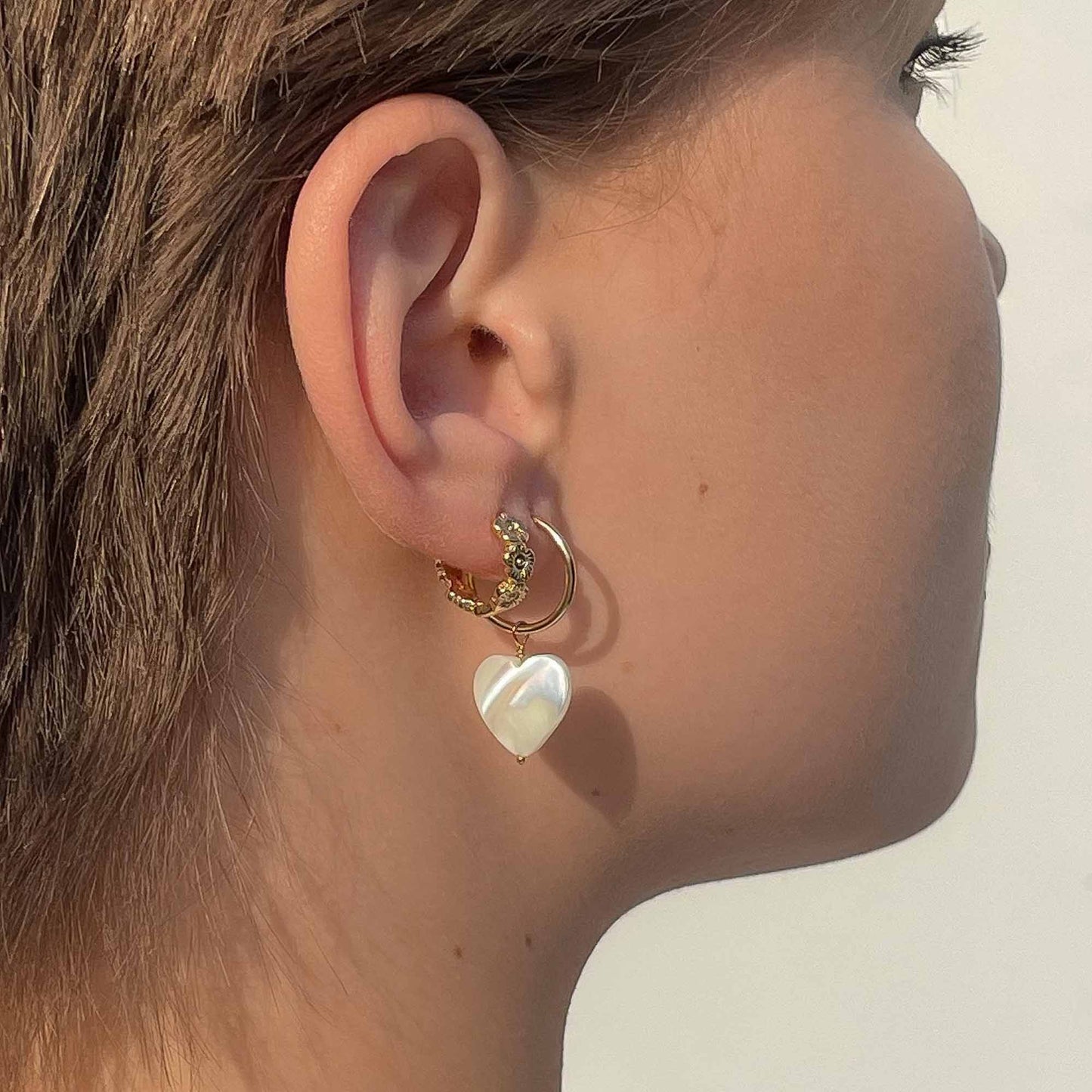 Close-up of an ear wearing a gold hoop earring with a heart-shaped pearl pendant on a neutral background.