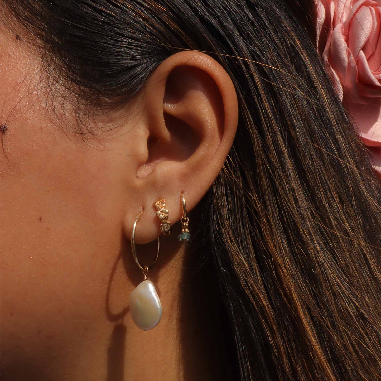 Close-up of an ear wearing gold hoop earrings with a pearl drop earring, pink flower in hair.