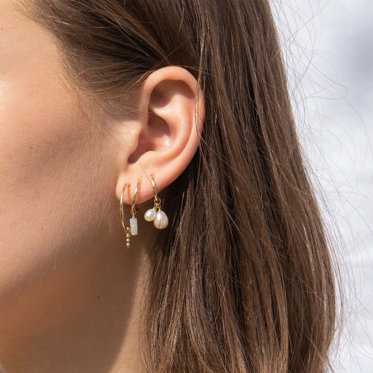 Close-up of an ear wearing gold hoop earrings with pearl-like beads against a neutral background.