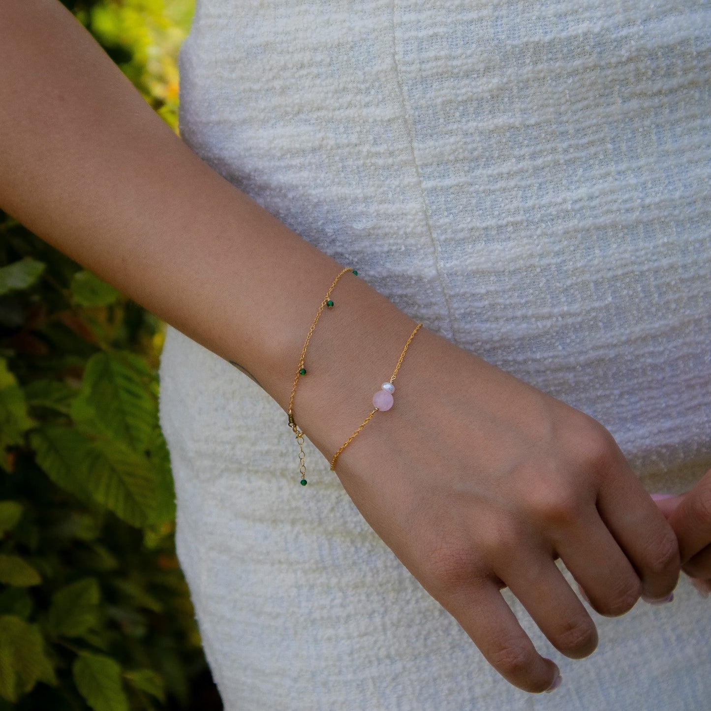 A close-up of a person's wrist wearing two delicate gold chain bracelets, one with tiny green beads and the other with a small pale pink stone, against a white textured dress and blurred green foliage background.