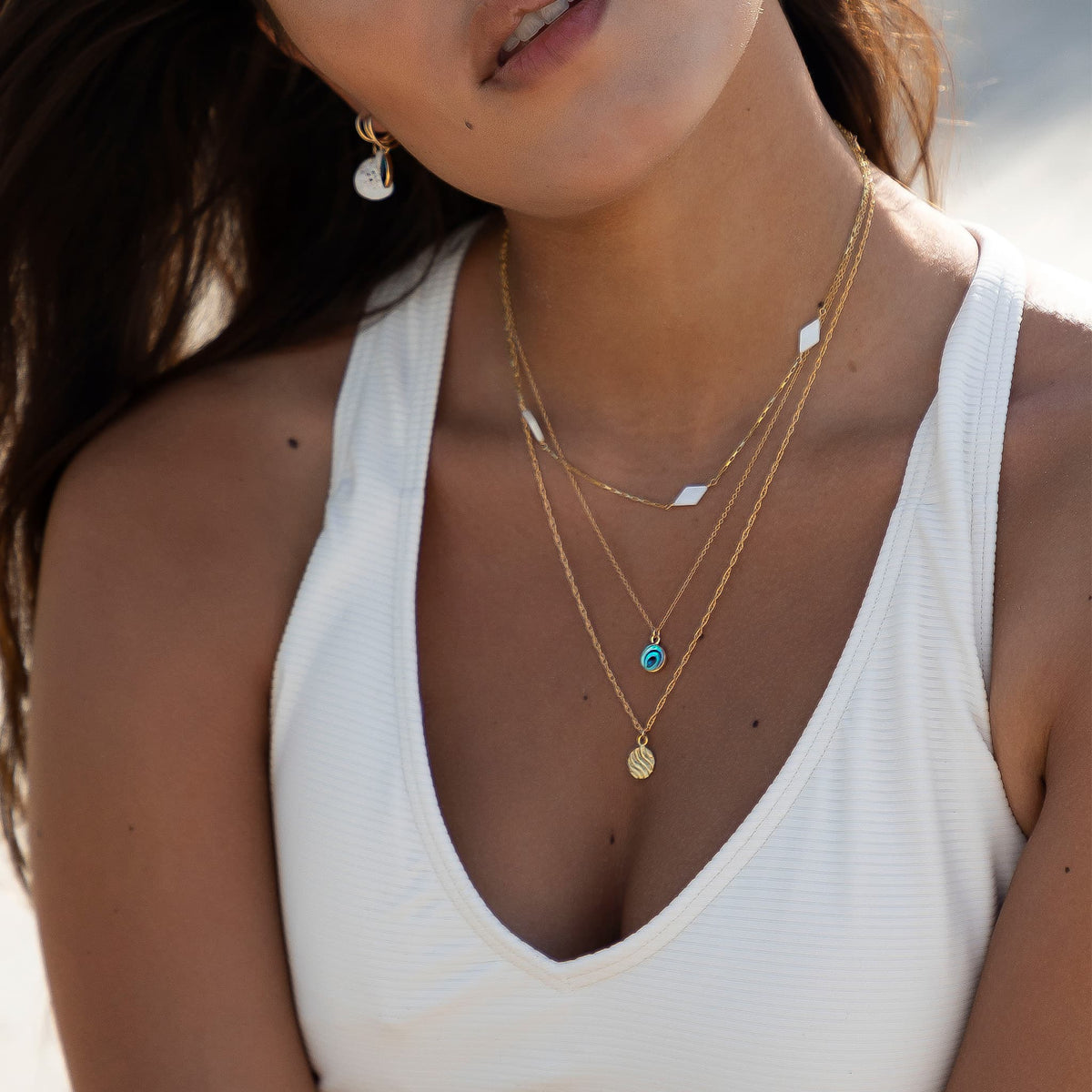 A close-up of a woman wearing layered gold necklaces, including a delicate chain with a small turquoise pendant and a textured gold disc charm, styled with a white ribbed tank top.