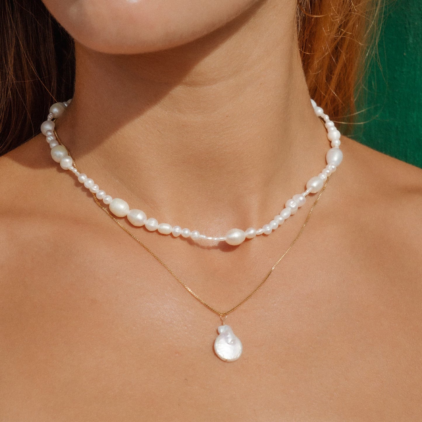 A close-up of a model wearing a double-layered necklace. The top layer is a short pearl choker, and the bottom layer is a delicate gold chain with a white teardrop-shaped pearl pendant.