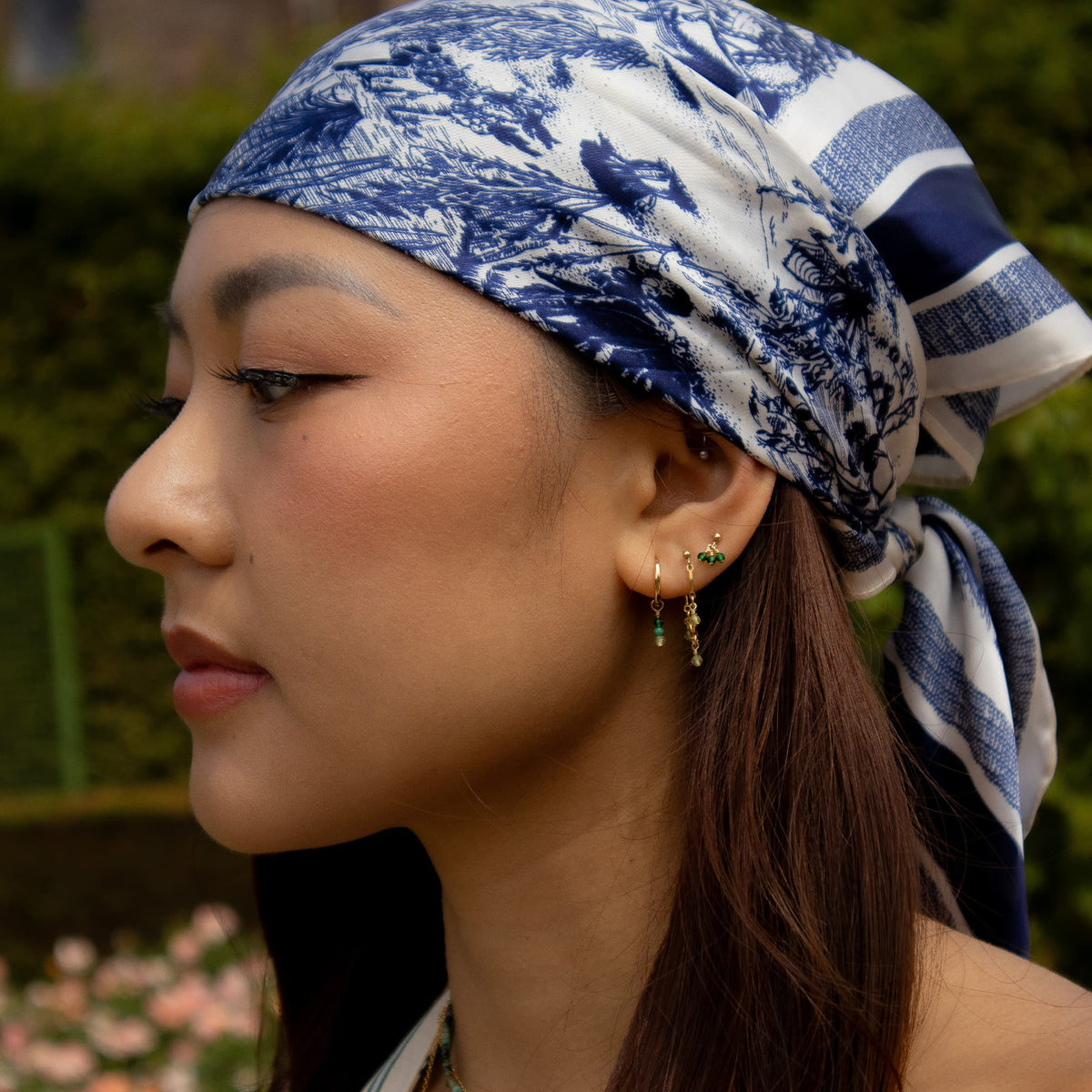 A close-up side profile of a woman wearing a blue and white toile-patterned headscarf and gold drop earrings with green stones, set against a blurred green garden background.