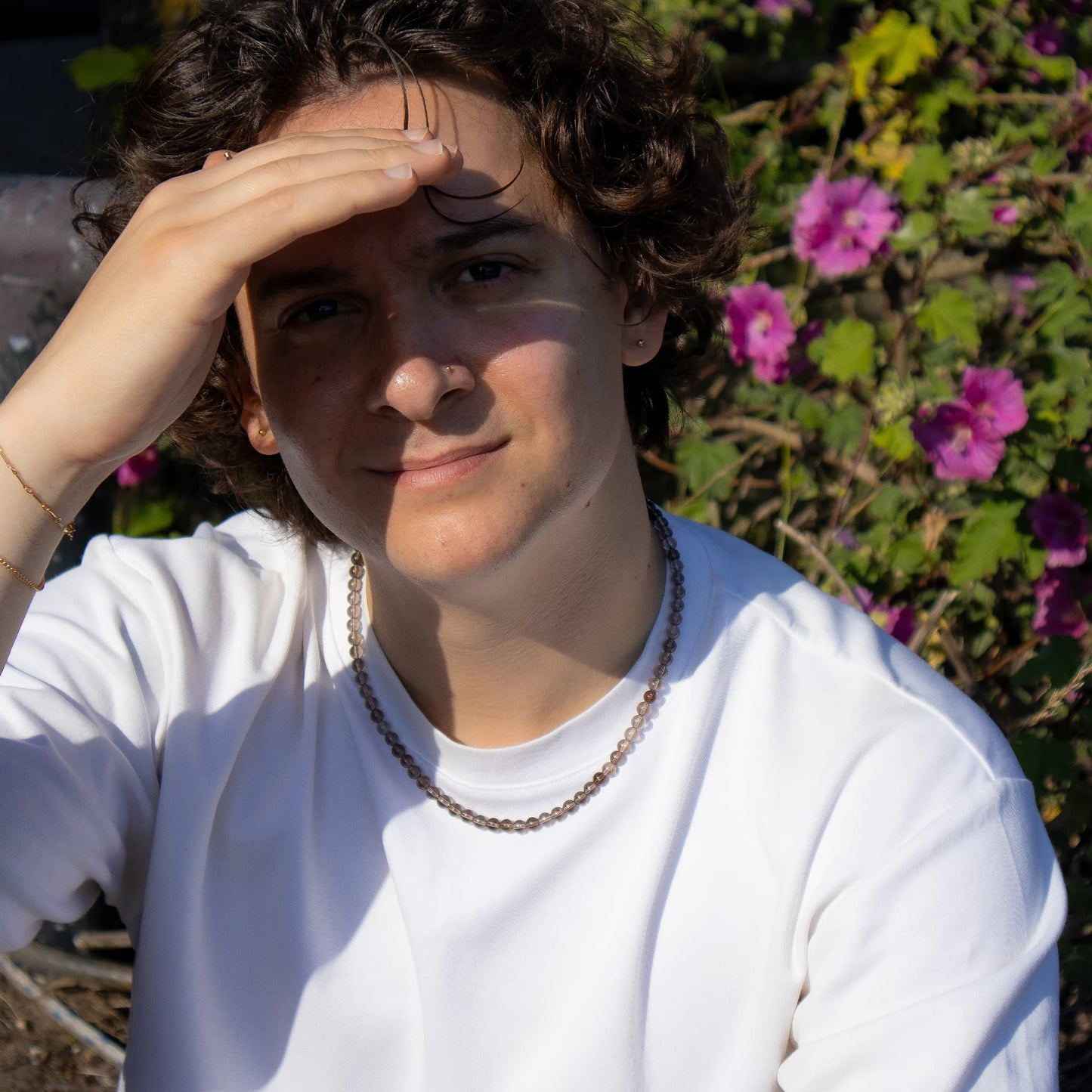 Person wearing a white shirt with a necklace, standing in front of pink flowers.