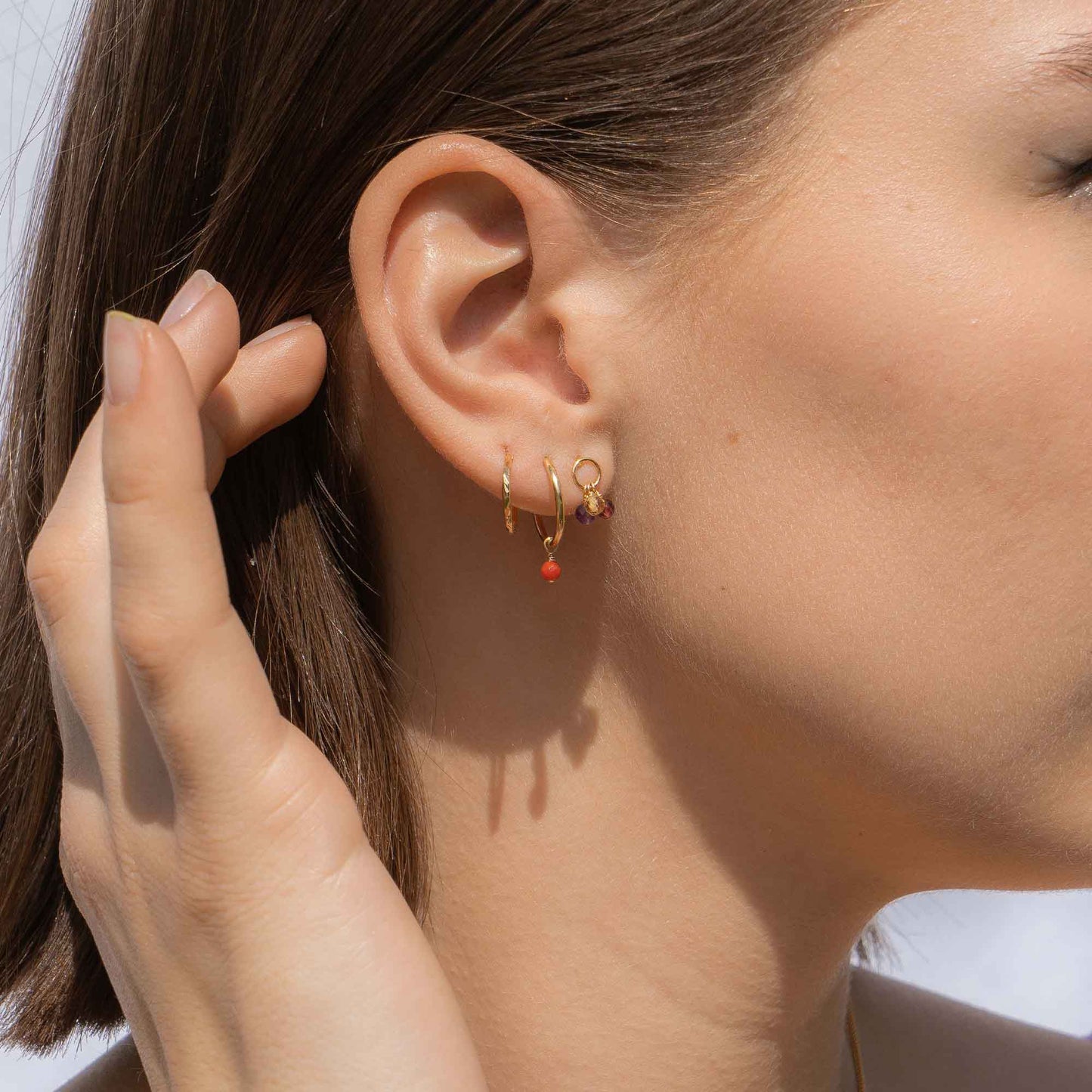 Close-up of an ear wearing gold hoop earrings with a red accent, with a hand adjusting the hair.