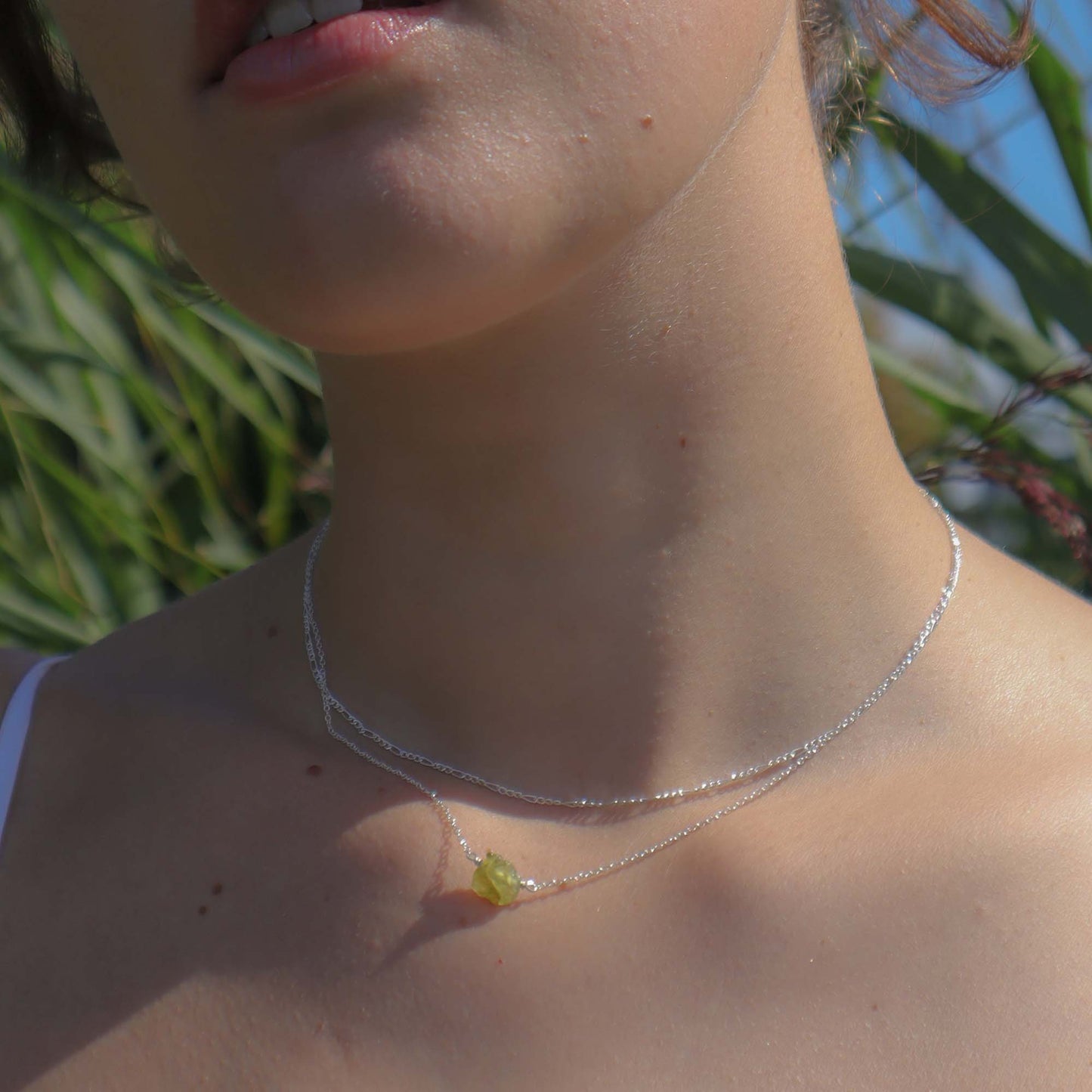 Close-up of a person wearing a silver necklace with a green pendant against a natural background.