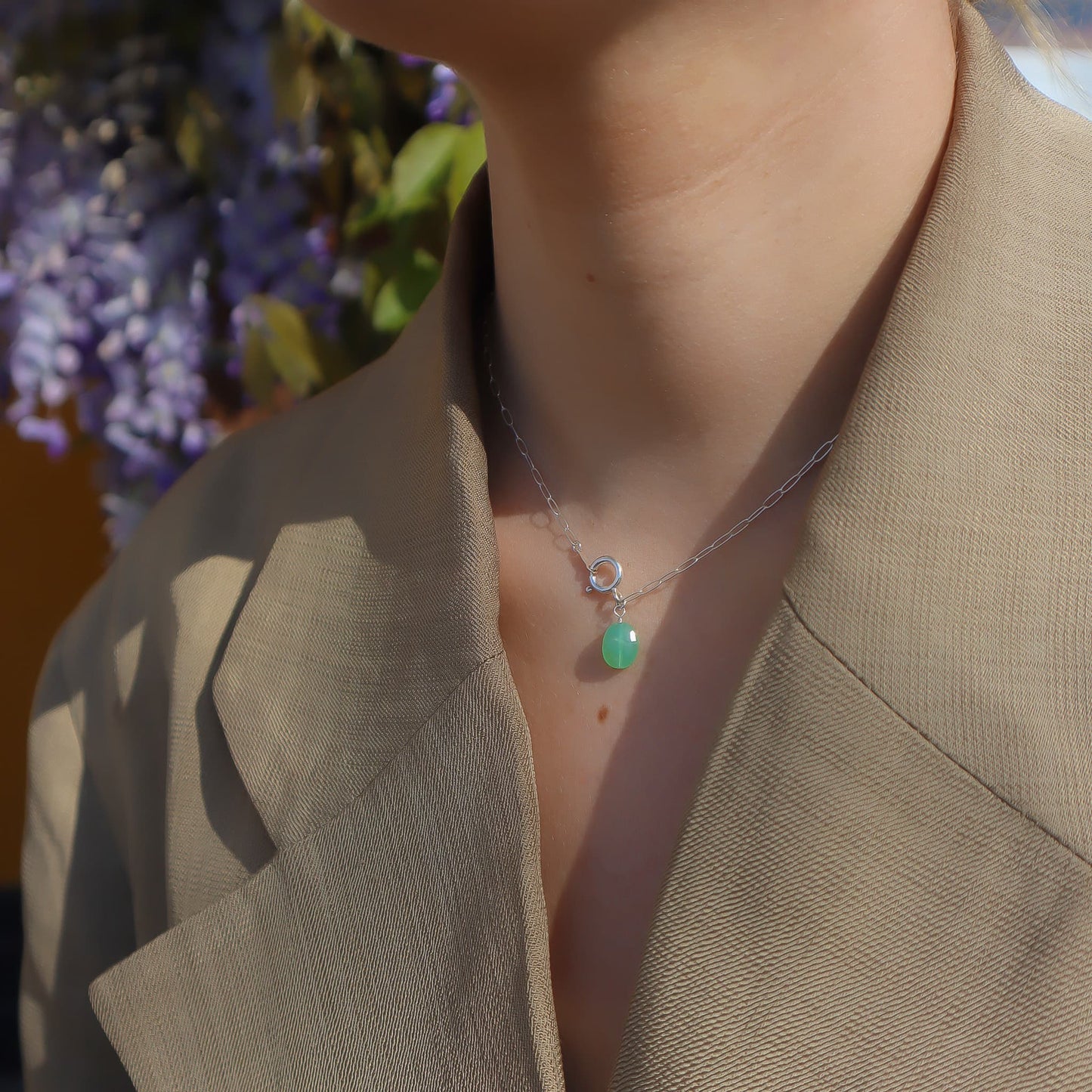 Necklace with a green pendant worn by a person in front of purple flowers