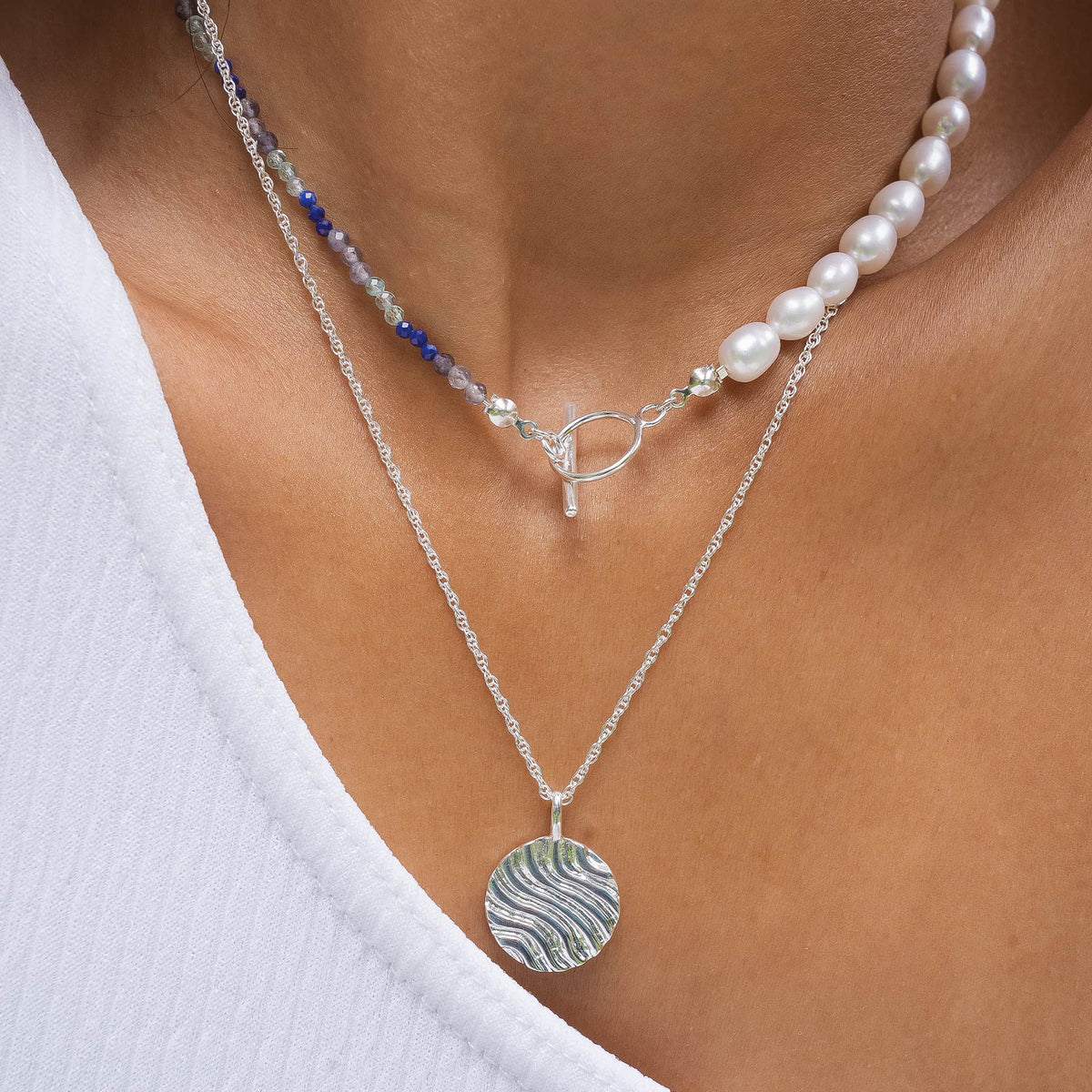 A close-up of a person's neck and chest wearing a layered necklace set. The set includes a silver chain with a round mother-of-pearl pendant featuring green and white wavy stripes, a pearl bead strand with a silver toggle clasp, and a delicate beaded strand with blue and clear beads.