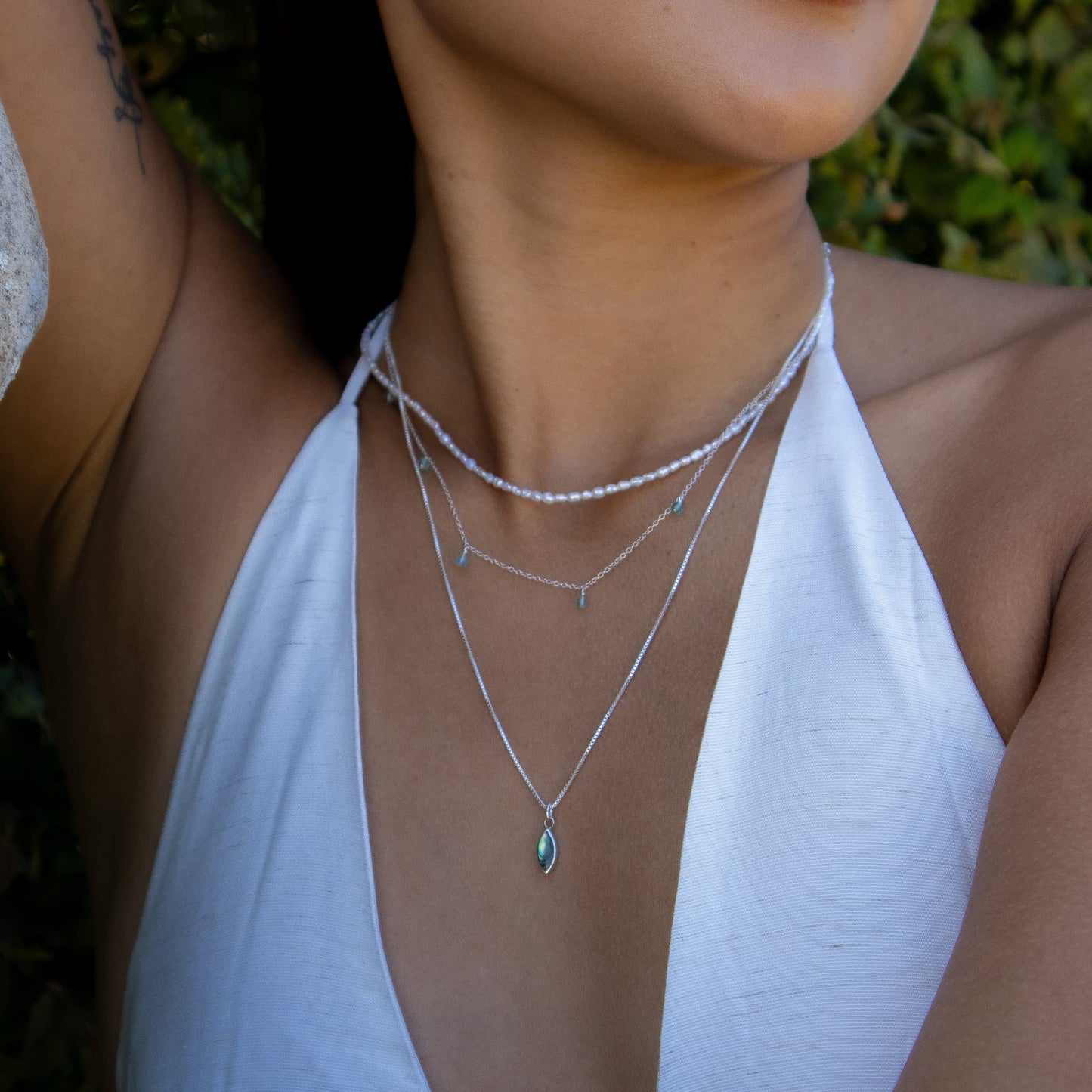A close-up of a woman wearing layered silver necklaces, including a beaded top layer, a chain with small dangling charms, and a longer chain with a teardrop-shaped green gemstone pendant, paired with a white halter top.