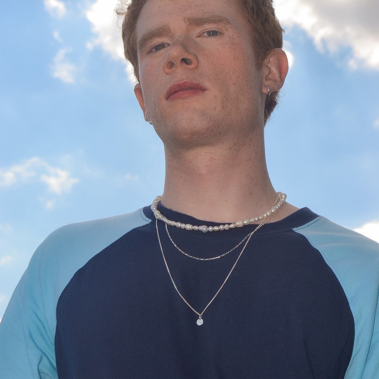 Person wearing a blue and black shirt with layered necklaces against a blue sky.