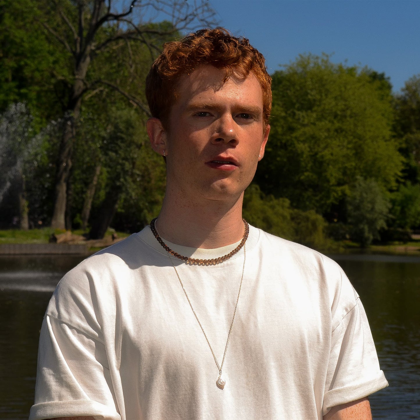 Person with red hair wearing a white shirt in a park setting with trees and water.