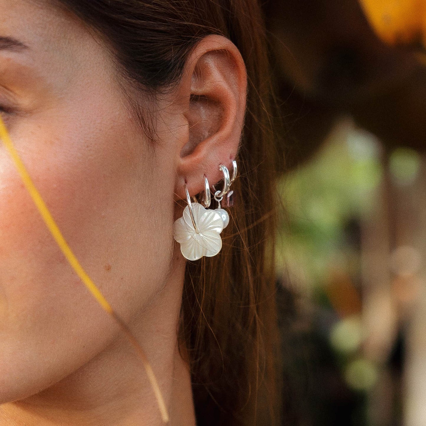 A close-up of a person's ear wearing a stack of silver hoop earrings with a white flower-shaped drop earring, set against a softly blurred outdoor background.
