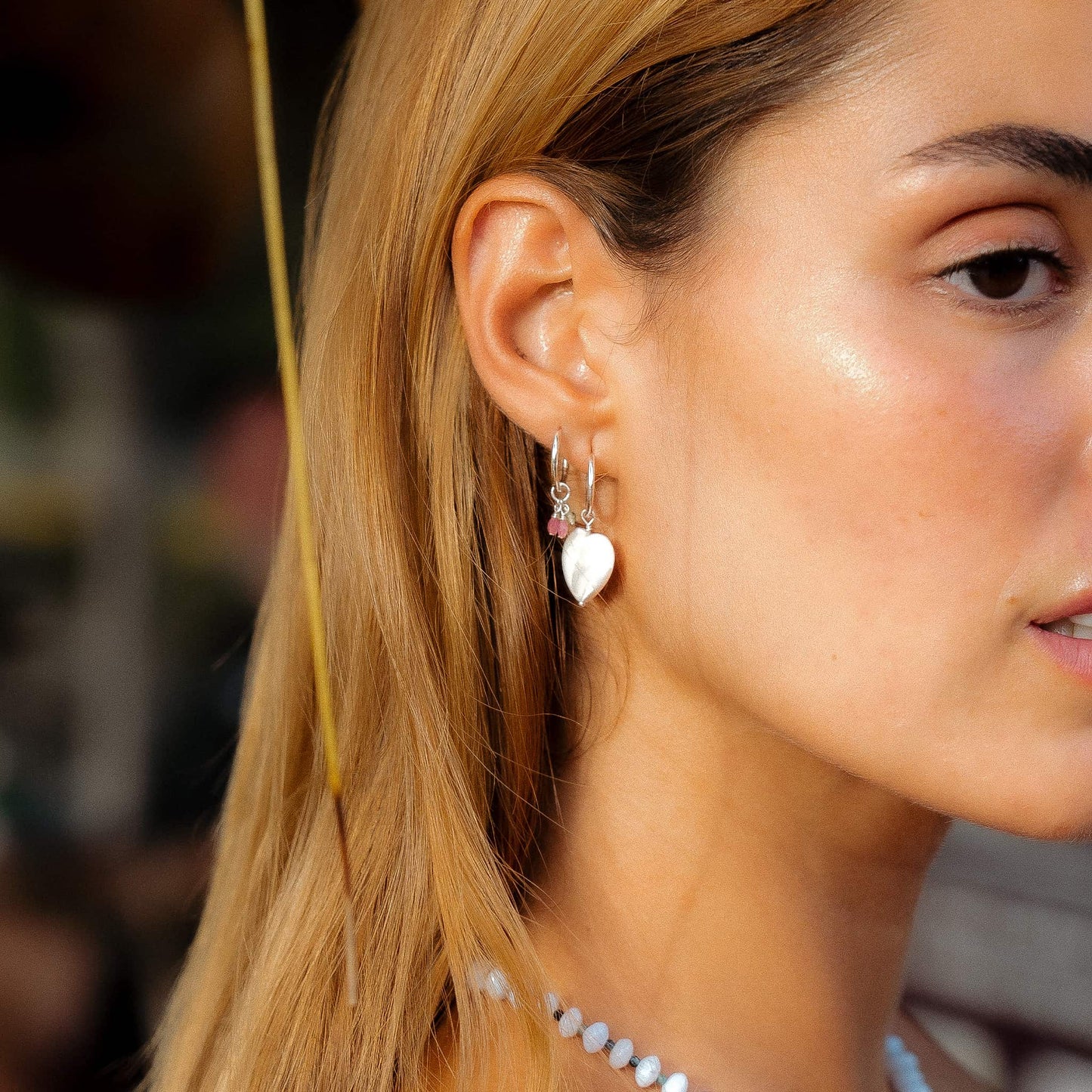 A close-up side profile of a woman with blonde hair wearing a silver hoop earring with a dangling white heart charm and small pink gem accents.